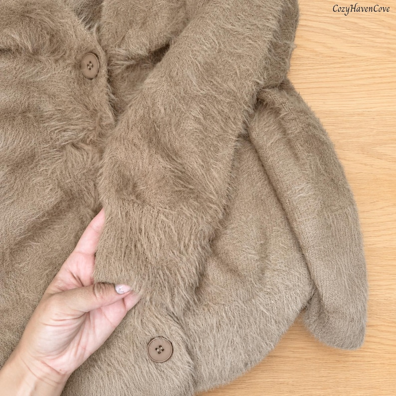 May include: A fuzzy, light brown cardigan with long sleeves and two visible round buttons. The knit fabric has a soft, textured appearance. The text CozyHavenCove is printed in the upper right corner of the image.