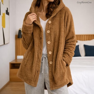 May include: A brown hooded coat with large wooden buttons. The coat has a soft, fuzzy texture and two front pockets. The text "CozyHavenCove" is in the upper right corner. The coat is worn over a white shirt and gray sweatpants.