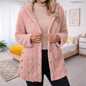May include: A light pink hooded coat with a textured, plush exterior. The coat features a button-down front with gold-colored buttons and long sleeves. The CozyHavenCove logo is visible in the upper right corner. The coat is worn over a light-colored turtleneck.