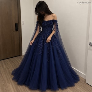 May include: A floor-length navy blue ball gown featuring an off-the-shoulder neckline, sheer tulle sleeves, and intricate floral lace appliques that cascade down the bodice and skirt. The voluminous skirt is crafted from layers of tulle fabric.