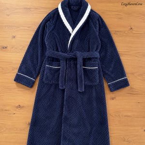 May include: A navy blue plush bathrobe with a textured waffle-knit pattern, featuring a white trim along the shawl collar, cuffs, and two front pockets. A matching tie belt is secured at the waist. The text CozyHavenCove appears in the corner.
