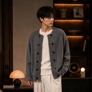 May include: A charcoal gray knit cardigan featuring a crew neckline, long sleeves, and a double-breasted button front closure. The garment is worn over a plain cream-colored sweater and light-colored trousers, showcasing a ribbed texture.