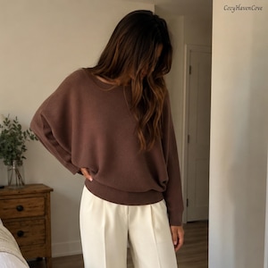 Könnte beinhalten: Ein brauner Langarmpullover mit weitem Ausschnitt und gerippten Bündchen, kombiniert mit einer cremefarbenen Hose. Der Pullover hat eine lockere Passform mit überschnittenen Schultern und ein strukturiertes Strickmuster. Der Text CozyHavenCove ist sichtbar.