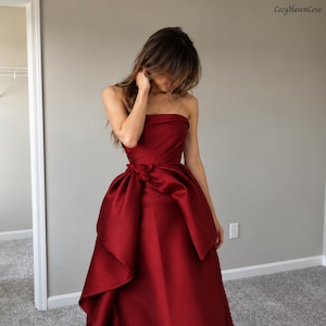 May include: A strapless floor-length gown in a deep burgundy red hue. The bodice features subtle diagonal pleating, while the waist is accented with a cluster of fabric flowers. The skirt has a voluminous, draped overlay that creates a layered effect.
