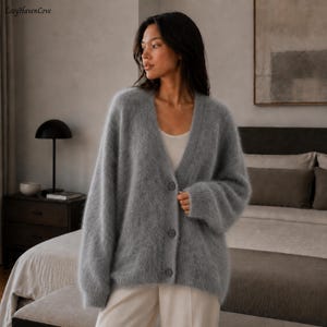 May include: A fuzzy, light gray V-neck cardigan featuring a button-down front with three matching gray buttons. The garment has long, loose sleeves and a soft, textured appearance. The text CozyHavenCove is visible in the upper left corner.