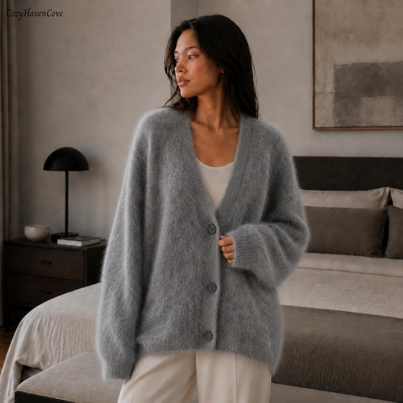 May include: A fuzzy, light gray V-neck cardigan featuring a button-down front with three matching gray buttons. The garment has long, loose sleeves and a soft, textured appearance. The text CozyHavenCove is visible in the upper left corner.