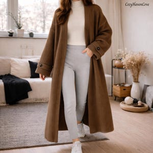 May include: A long, brown ribbed knit cardigan with buttoned cuffs. The open-front sweater is paired with grey trousers and white trainers. The text "CozyHavenCove" is visible in the upper right corner. The overall theme is casual wear.