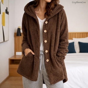 May include: A brown hooded jacket with large wooden buttons and pockets. The jacket is made of a soft, plush material. The CozyHavenCove logo is in the upper right corner. The person is wearing a white shirt and gray sweatpants.