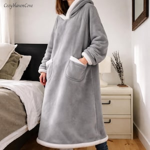 Può includere: Una coperta con cappuccio grigia con finiture e tasche bianche. La coperta è lunga, arriva alle caviglie, e ha una consistenza morbida e soffice. Il cappuccio e le tasche sono foderati in tessuto bianco. Il marchio "CozyHavenCove" è visibile in alto a sinistra.