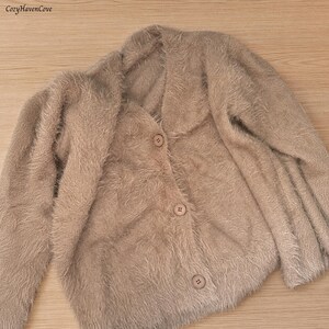May include: A beige fuzzy cardigan with long sleeves, a V-neckline, and three matching buttons down the front. The garment features a soft, textured, shaggy knit construction. The text CozyHavenCove is visible in the upper left corner.