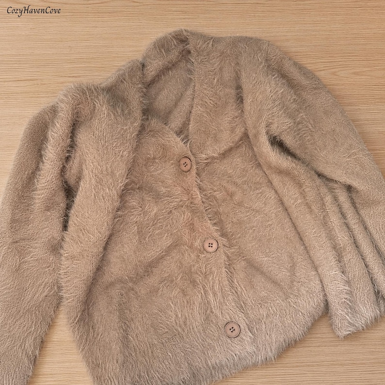May include: A beige fuzzy cardigan with long sleeves, a V-neckline, and three matching buttons down the front. The garment features a soft, textured, shaggy knit construction. The text CozyHavenCove is visible in the upper left corner.