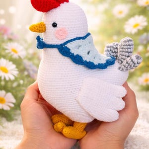 Puede incluir: Un pollo blanco de ganchillo hecho a mano con una cresta roja, pico y patas amarillas y una bufanda azul. El pollo tiene una mejilla rosa y alas grises y blancas. El pollo se sostiene con dos manos.