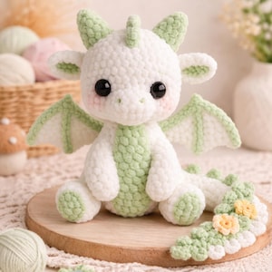 Puede incluir: Un dragón de ganchillo blanco y verde hecho a mano, con grandes ojos negros, sentado sobre un círculo de madera. El dragón tiene alas y detalles verdes. La cola está decorada con pequeñas flores amarillas. El fondo incluye hilo.