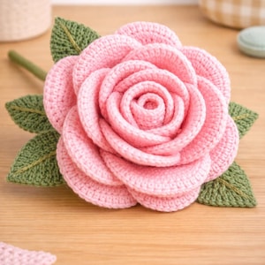 Puede incluir: Una gran rosa de ganchillo hecha a mano en rosa suave, con hojas y tallo verdes. Los pétalos están meticulosamente elaborados, mostrando la textura del hilo. Esta pieza floral decorativa es perfecta para la decoración del hogar o como un regalo único.