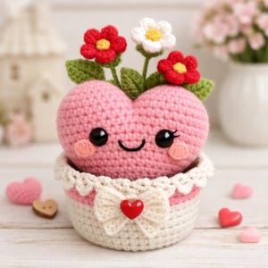 Puede incluir: Una figura en forma de corazón rosa de ganchillo con una cara sonriente, rubor y ojos negros grandes, se encuentra en una cesta de ganchillo blanca y rosa. El corazón tiene hojas verdes y flores de ganchillo rojas y blancas. Un lazo blanco con un corazón rojo está en la cesta.