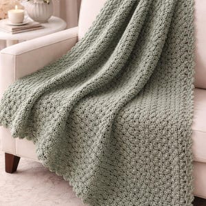 Puede incluir: Una manta de ganchillo verde salvia sobre un sofá blanco. La manta tiene un patrón texturizado en relieve y un borde festoneado. El sofá está en una habitación bien iluminada con un jarrón de flores y una vela en una mesa auxiliar.