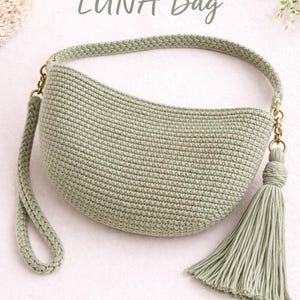 Puede incluir: Un bolso de ganchillo verde salvia con forma de media luna, un asa trenzada y una borla. El bolso tiene herrajes dorados y se muestra el texto "LUNA Bag". También es visible el texto "Easy Pattern • Advanced Beginner".