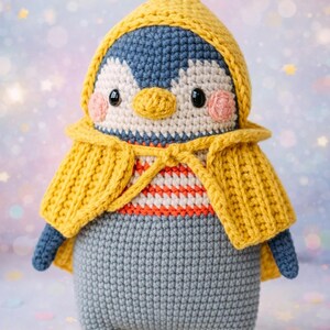 May include: Handmade crochet penguin doll wearing a yellow hooded cape and a striped shirt. The penguin has a blue and white head, pink cheeks, and gray pants. The doll is approximately 8 inches tall.
