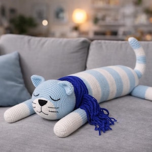 Könnte beinhalten: Ein handgefertigtes gehäkeltes Katzenplüschtier in hellblauen und weißen Streifen, mit blauem Kopf und blauem Schal. Die Katze hat geschlossene Augen und weiße Pfoten. Das Plüschtier liegt auf einem grauen Sofa.