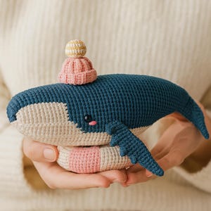 May include: A handmade crochet whale in shades of blue, cream, and pink, with a small pink and yellow hat. The whale has a friendly expression and is being held in someone's hands.