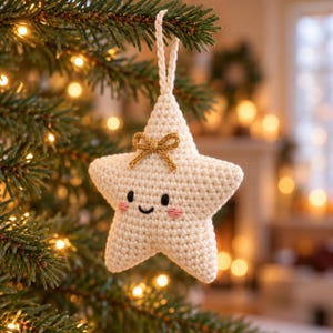 Puede incluir: Un adorno de estrella de ganchillo color crema con un pequeño lazo dorado en la parte superior, ojos y boca sonriente bordados en negro, y mejillas cosidas en rosa. La estrella incluye un lazo de ganchillo a juego para colgarla en el árbol.