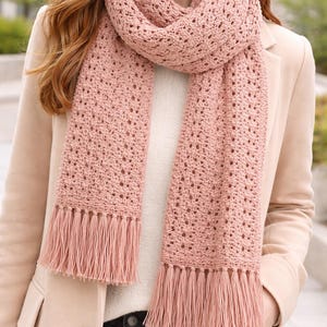Puede incluir: Una bufanda rosa claro de tejido abierto con flecos. La bufanda está hecha de un hilo suave y tiene un delicado diseño calado. La bufanda se lleva alrededor del cuello y los hombros, y se combina con un suéter crema y una blazer beige claro.