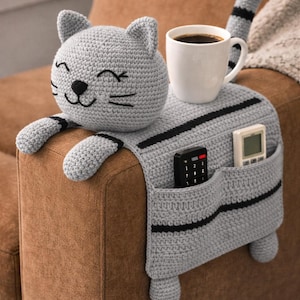 May include: A gray crocheted cat-shaped couch armrest organizer with black stripes and pockets. The cat has a smiling face and paws. A white mug of coffee and two remote controls are in the pockets.