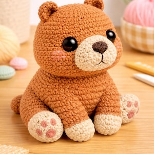 Puede incluir: Un oso de peluche de ganchillo hecho a mano en tonos marrón y crema. El oso tiene grandes ojos negros, un hocico color crema y mejillas rosadas. Las patas presentan almohadillas rosas. Este juguete blando es perfecto para una guardería o como regalo.
