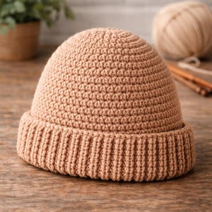 Op de afbeelding: Een bruin gehaakte beanie met een geribbelde rand. De hoed is gemaakt van een zacht, getextureerd garen en ligt op een houten oppervlak. Een bol garen en breinaalden staan op de achtergrond.