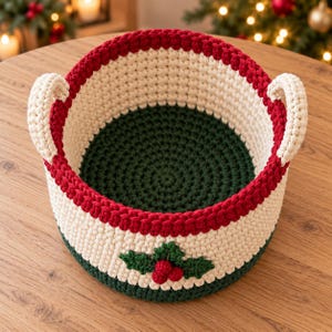 Puede incluir: Una cesta redonda de ganchillo con base e interior verde oscuro, una sección central color crema y un borde rojo vibrante. Dos asas color crema están unidas a los lados. Un motivo de hojas de acebo y bayas de ganchillo está centrado.