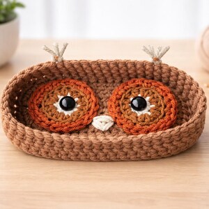 May include: A handmade crochet owl basket in shades of brown, orange, and white. The basket features large, round eyes with black pupils and a small white beak. Two small tufts of yarn are attached to the top. This decorative item is perfect for home decor.