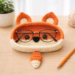 May include: A crocheted fox-shaped glasses holder in orange and cream colors. The holder has a fox face design with black button eyes, a black heart-shaped nose, and two ears. A pair of glasses rests inside the holder, and a small fox tail is next to it.