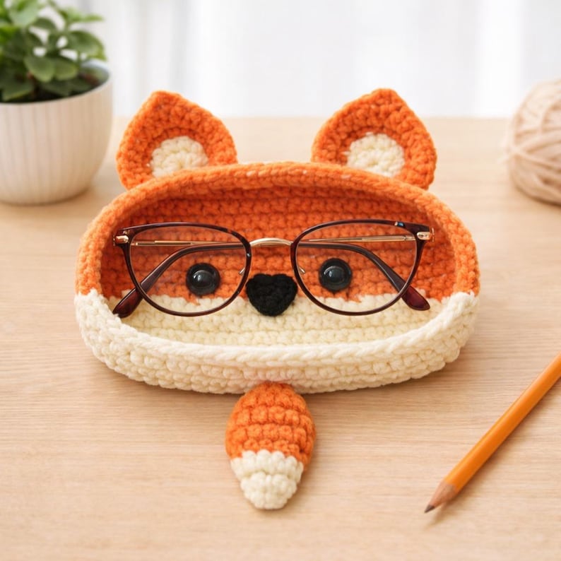 May include: A crocheted fox-shaped glasses holder in orange and cream colors. The holder has a fox face design with black button eyes, a black heart-shaped nose, and two ears. A pair of glasses rests inside the holder, and a small fox tail is next to it.