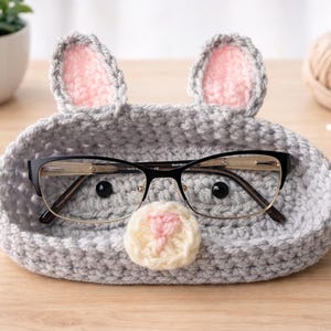 May include: A gray crocheted eyeglass holder shaped like a mouse, with pink-lined ears and a cream-colored nose. Black-framed glasses rest inside the holder. The holder is on a wooden surface, with a small plant and yarn in the background.