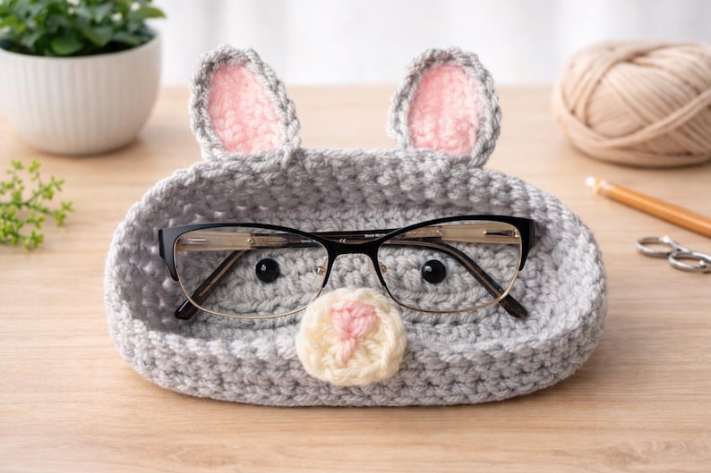 May include: A gray crocheted eyeglass holder shaped like a mouse, with pink-lined ears and a cream-colored nose. Black-framed glasses rest inside the holder. The holder is on a wooden surface, with a small plant and yarn in the background.