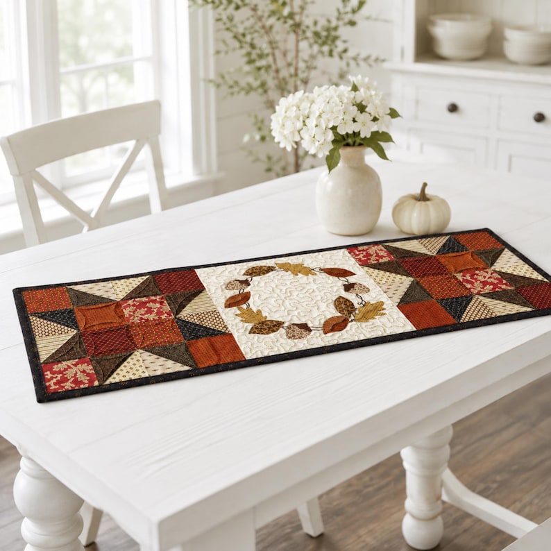 May include: A rectangular quilted table runner featuring a central panel with an embroidered autumn leaf wreath on a cream background, flanked by two square blocks with geometric star patterns in shades of rust, brown, tan, and black.