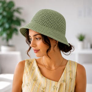 May include: A sage green crochet bucket hat featuring a textured, woven stitch pattern with a rounded crown and a downward-sloping brim. The hat is constructed from a single solid color yarn, showcasing a consistent, dense, and uniform knit design.