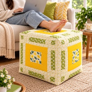 May include: A square fabric ottoman featuring a patchwork design with bright yellow solid panels, white fabric with small yellow flower prints, and green fabric with white daisy patterns, all stitched together with a visible quilted grid texture.
