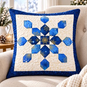 May include: A square throw cushion featuring a central quilt-style snowflake pattern. The design uses shades of blue and cream fabric with gold snowflake motifs. The cushion has a dark blue border and a cream-coloured background with quilted texture.