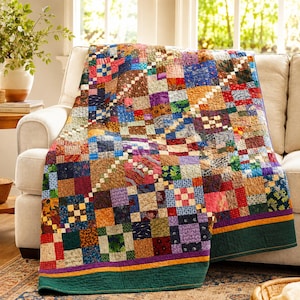 May include: A patchwork quilt featuring a grid of multicolored fabric squares in various patterns including floral, plaid, and geometric prints. The quilt has a wide dark green border at the bottom edge, accented by a thin purple and orange stripe.