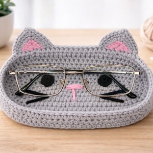 May include: A gray crocheted cat-shaped tray with pink ear accents and a pink nose. The tray holds a pair of eyeglasses with a silver frame. The tray is on a wooden surface.