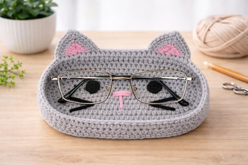 May include: A gray crocheted cat-shaped tray with pink ear accents and a pink nose. The tray holds a pair of eyeglasses with a silver frame. The tray is on a wooden surface.