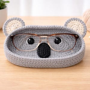 May include: A handmade gray crochet koala-shaped tray with a black nose and two black eyes, holding a pair of eyeglasses. The tray has two beige ears. A decorative item for glasses or small items.