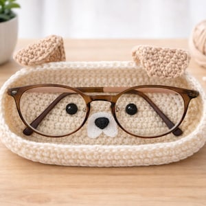 May include: A cream-colored, crocheted eyeglasses holder shaped like a dog, with brown-framed glasses inside. The holder has two beige ears and a felt nose. The glasses have a brown frame. The holder is on a wooden surface.