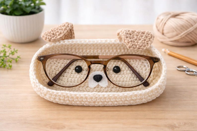 May include: A cream-colored, crocheted eyeglasses holder shaped like a dog, with brown-framed glasses inside. The holder has two beige ears and a felt nose. The glasses have a brown frame. The holder is on a wooden surface.