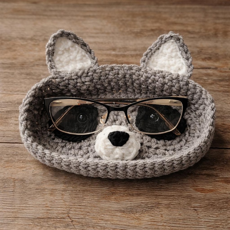 May include: A gray crocheted raccoon-shaped glasses holder with white inner ears and a black and white nose. The holder contains a pair of black-framed eyeglasses. The holder is sitting on a wooden surface.