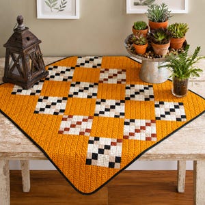 May include: A square quilted table topper featuring a checkerboard pattern of solid mustard yellow squares and pieced squares containing smaller white, tan, and black squares. The quilt has a thin black border and rounded corners.