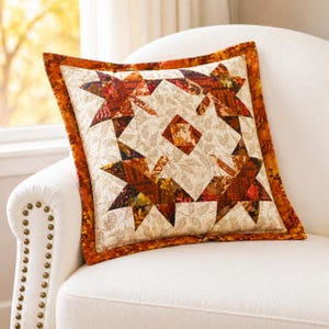 May include: A square cushion with a patchwork design. The cushion features a central pattern of four star shapes in shades of orange, brown, and yellow, set against a beige background with a leaf pattern. The border is a solid orange.