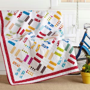 Puede incluir: Una colcha blanca con un diseño de patchwork de tiras rectangulares de tela en varios colores, incluyendo rojo, amarillo, azul, verde y marrón, con patrones florales y de lunares. La colcha tiene un borde rojo sólido y costuras visibles.