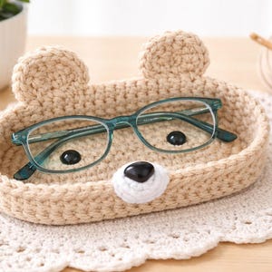 May include: A handmade, beige crochet bear-shaped tray holding a pair of teal-framed eyeglasses. The tray has two round ears and a white nose. The tray sits on a white, lace doily. A ball of yarn and a crochet hook are in the background.